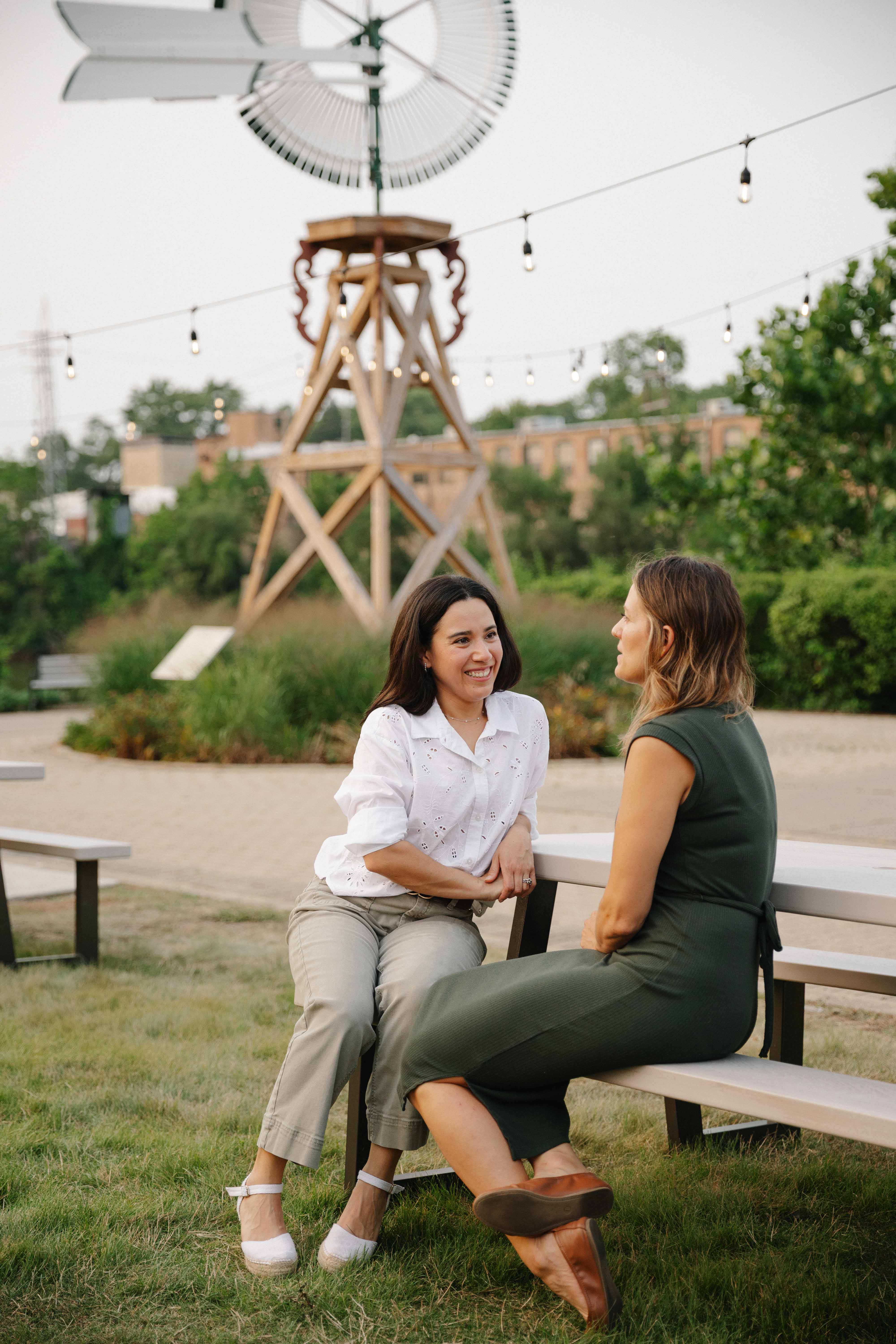 Maria and Sarah talking at a picnic table in downtown Batavia, under the windmill and string lights